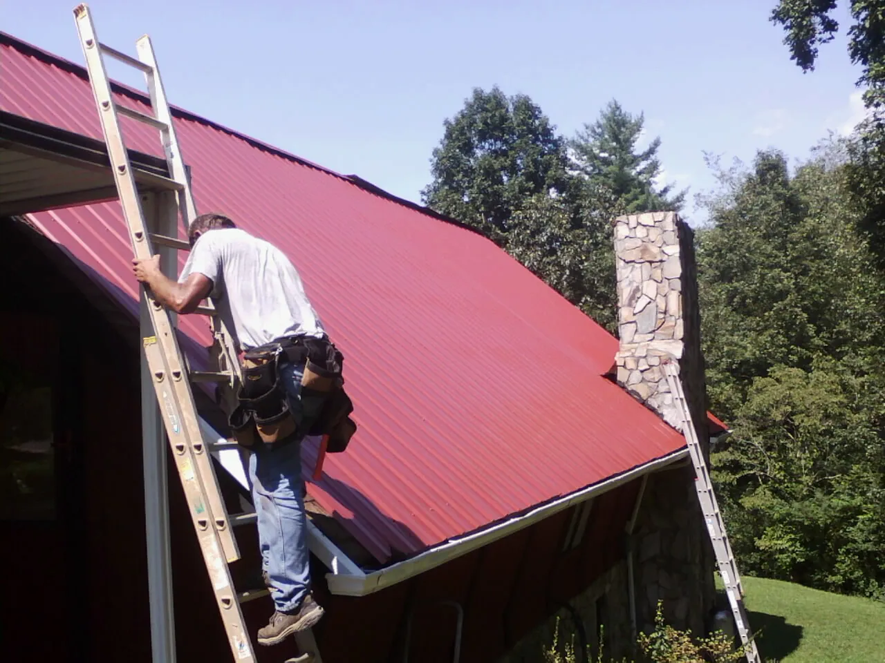 Roofer on a ladder beside a red metal roof and stone chimney — Soffit & Fascia Repair by Roofing Brothers in Center Valley